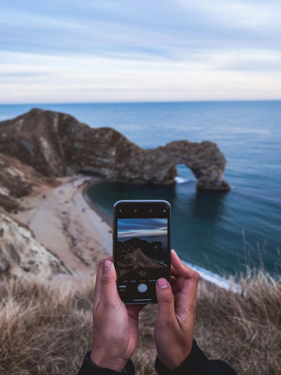 Joku henkilö ottamassa maisemakuvaa älypuhelimella rantakielekkeen päältä. Kuvassa näkyy lisäksi pieni ranta, meri ja kallioita.